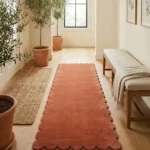Perspective lifestyle view of the Autumn Embrace runner rug placed on a warm wooden floor in a brightly lit entryway decorated with potted plants and a bench, featuring a solid warm terracotta orange color and a continuous contoured scalloped edge.