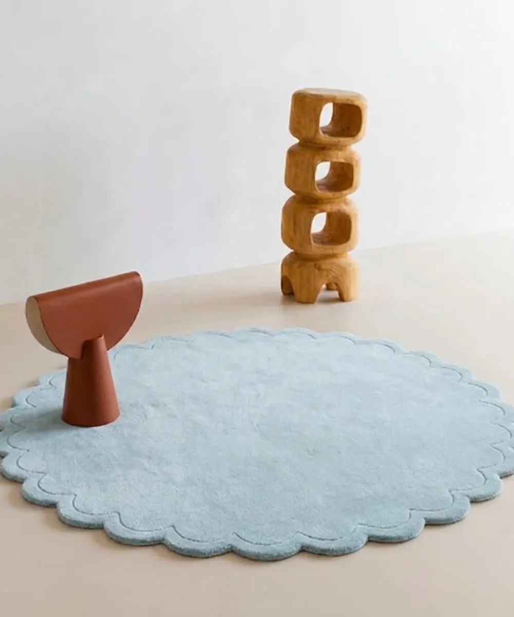 Ice Whisper circular blue rug with scalloped edges styled in a bright modern room with wooden art pieces.