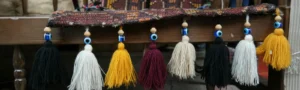 A close-up banner image of a textured woven fabric edge decorated with hanging yarn tassels in alternating colors of black, white, golden yellow, and burgundy, with each tassel topped by a classic blue glass evil eye bead.