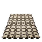Low angle perspective view of the Heritage Lattice rug, featuring a repeating geometric chain-link pattern in dark brown on a soft beige background, framed by a continuous dark brown scalloped edge.