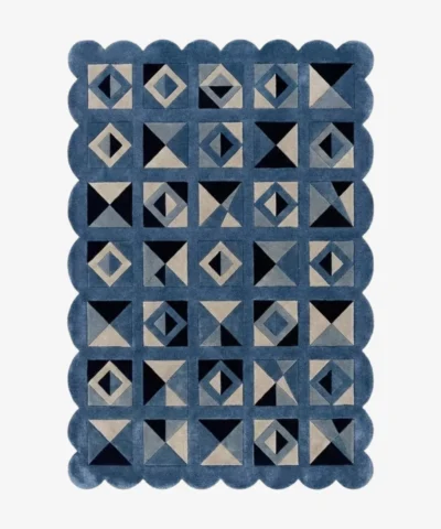 Top down view of the Indigo Tessellate rug against a white background, featuring a repeating patchwork pattern of geometric squares, triangles, and diamonds in solid shades of indigo blue, grey, and black, framed by a continuous blue scalloped border.