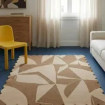 Perspective view of the Sand Fragments rug styled in a modern room with a yellow chair, featuring abstract geometric shapes in warm beige and cream tones.