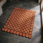 Perspective view of the Terracotta Grid rug placed on a dark tiled floor, showcasing the bold repeating geometric diamond lattice design and the structured scalloped border illuminated by natural sunlight.