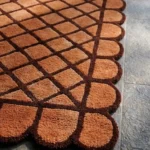 Close-up angled view of the Terracotta Grid rug, highlighting the plush hand-tufted New Zealand wool and viscose silk texture, the deeply carved dark brown lines defining the geometric diamond lattice pattern against the warm terracotta background, and the intricately contoured scalloped edge.