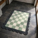 High angle view of the Lune Mist rug featuring a soft sage green and cream checkerboard pattern, framed by a dark charcoal grey scalloped border.