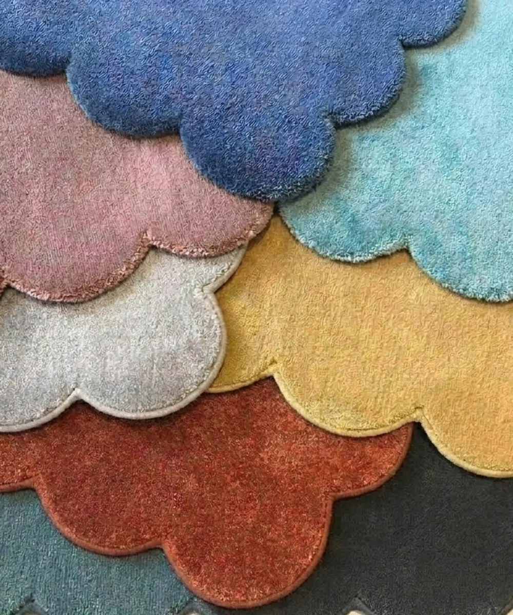 Close-up top-down view of a stacked collection of solid color scalloped rugs in various shades including blue, pink, yellow, and red, highlighting the thick hand-tufted texture for the shop all rugs category.