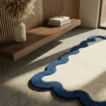 A close-up, angled view of the Azure Wave Scallop Rug resting on a light concrete floor. A beam of sunlight emphasizes the thick, tufted yarn texture of the cream center and the contrasting dark blue wavy border.