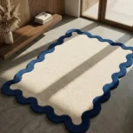 An angled shot of the Azure Wave Scallop Rug placed on a smooth concrete floor in a sunlit, minimalist room. The rug's plush cream center and distinct blue scalloped edges are highlighted by natural light next to a wooden slat wall and shelf.