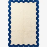 A top-down, flat view of the Azure Wave Scallop Rug on a clean white background. The tufted rug features a large rectangular cream center bordered by a thick, wavy, dark blue scalloped edge.