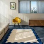 The rectangular Azure Wave Scallop Rug styled in a bright, modern living room with a view of the city. The cream tufted rug with its bold blue scalloped border sits on a blue floor near a yellow accent chair, a light sofa, and a wooden dresser.