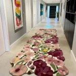 A long perspective view of the Cassia Floral Runner Rug placed down the center of a modern, brightly lit hallway with light tiled floors. The thick tufted rug adds a vibrant pop of pink, burgundy, and green botanical patterns to the neutral corridor.
