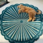 A fluffy golden doodle dog sleeping peacefully on the circular Emerald Sunburst Round Rug. The bright sunlight highlights the rug's plush tufted texture and its radiating teal, emerald green, and cream geometric pattern.