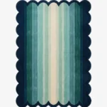 A flat, top-down view of the rectangular Ocean Drift Scallop Rug on a clean white background. The tufted rug features a wavy, dark blue scalloped border and a vertical striped gradient transitioning from dark teal on the outside to light mint green and a cream center.