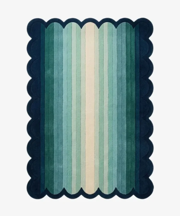 A flat, top-down view of the rectangular Ocean Drift Scallop Rug on a clean white background. The tufted rug features a wavy, dark blue scalloped border and a vertical striped gradient transitioning from dark teal on the outside to light mint green and a cream center.