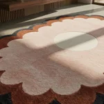 A close-up, angled view of the Sunburst Flower Hand Tufted Wool Rug resting on a smooth concrete floor in a sunlit room. The image highlights the thick, plush yarn texture of the light cream center and the surrounding layered scalloped petals in soft pink, warm rust orange, and dark grey.