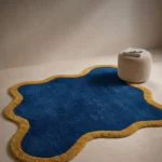 Custom-shaped Indigo Tide rug with a vibrant blue center and solid mustard yellow border styled on a neutral floor next to a modern round pouf.