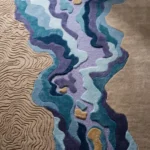 A detailed close-up of the Lavelle Drift Rug highlighting its incredible hand-tufted craftsmanship. The image captures the dynamic 3D textures of the deeply carved beige topographic lines, contrasting beautifully with the plush, multi-level New Zealand wool and viscose yarn that forms the flowing blue and purple abstract water design.