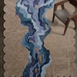 An overhead angled view of the Lavelle Drift Rug resting on a dark concrete floor. The modern scalloped rug showcases its intricate beige topographic carving and vibrant blue and purple river pattern, beautifully complemented by a mid-century modern wooden chair placed partially on its soft, plush surface.