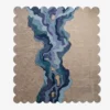 A flat, top-down view of the Lavelle Drift Rug isolated on a clean white background. The hand-tufted scalloped rug features a unique landscape-inspired design, showcasing carved topographical lines on a sandy beige base, intersected by a flowing, multi-colored blue and deep purple abstract river motif.