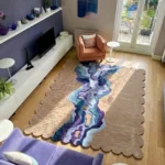 An overhead view of the Lavelle Drift Rug styled in a bright, contemporary living room. The custom scalloped edge rug rests on a light wood floor, bathed in natural sunlight, and is beautifully positioned between a plush purple sofa, an orange armchair, and a sleek white media console to highlight its striking organic design.