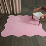 An overhead angled view of the pink Roselle Rug styled in a modern room setting. The beautifully shaped scalloped rug rests on a smooth dark concrete floor next to flowing beige curtains, with a mid-century modern wooden chair featuring a white seat placed partially on its soft surface.