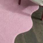 An extreme close-up of the rose pink Roselle Rug gently folded over itself. The detailed shot highlights the elegantly carved inner wavy border and the thick, plush tufted yarn construction. The image emphasizes the rug's high-quality internal stuffing designed for luxurious, natural softness.