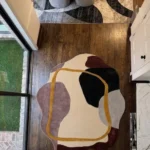 An overhead view of the abstract Terra Frame Scallop Rug placed on dark hardwood floors in a modern home hallway. The tufted rug's organic shape and contemporary color-blocking contrast beautifully with the rich wood floor, positioned right next to sliding glass doors that look out onto green grass.
