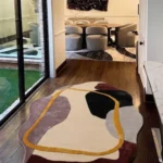 An angled view of the hand-tufted Terra Frame Scallop Rug resting on a dark hardwood hallway floor. The modern, organic-shaped rug showcases a cream background with black, grey, mauve, and dark red abstract shapes, highlighted by a yellow continuous line frame, perfectly styling the walkway leading into a warmly lit, contemporary dining room.