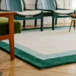 Low-angle perspective shot showing the Avelis border rug placement under green wooden chairs on a herringbone floor.
