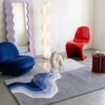 Wide perspective interior lifestyle view of the Coastal Meridian abstract landscape rug styled on a grey concrete floor in a modern room with an acrylic coffee table and colorful accent furniture.