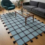 Wide perspective interior lifestyle shot of the light blue Crosshatch grid pattern rug styled on a hardwood floor in a mid-century modern living room with a grey sofa and teal accent chair.