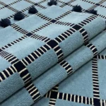 Close-up macro photograph highlighting the premium wool material texture, dense pile tracking, and clean striped grid work of the folded Crosshatch blue rug.