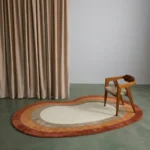 Wide perspective interior lifestyle shot of the irregular Elio Bend kidney rug styled on a green concrete floor next to a wooden mid-century modern chair and floor-to-ceiling curtains.