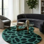Wide perspective interior lifestyle view of the kidney shaped Fernfall green rug styled in a modern living room under a glass coffee table with a dark curved sofa.