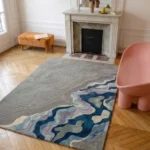 Wide perspective interior lifestyle view of the Gray Meridian topographic rug styled on a classic herringbone hardwood floor in an elegant room featuring a white fireplace mantel and a contemporary pink statement chair.
