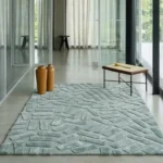 Wide perspective interior lifestyle shot of the Mist Glacis geometric block rug styled on a smooth concrete floor in a minimalist room with glass partition walls and modern accent furniture.