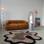 Mocha Drift custom organic rug styled flat on a concrete floor in a modern interior setting featuring an ochre brown couch, a wavy floor lamp, and a purple accent stool.