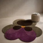 Wide perspective interior lifestyle shot of the orchid-shaped Plum Orchid accent rug styled on a minimalist concrete floor next to a neutral pouf ottoman.
