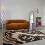 Wide perspective interior lifestyle shot of the Shadow Canyon rug styled on a concrete floor in a modern living room with an orange sofa and eclectic decor.