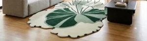 An interior lifestyle photograph of an irregular, organic-shaped area rug with a green and cream botanical pattern, styled on light hardwood flooring between a modern sofa and coffee table.