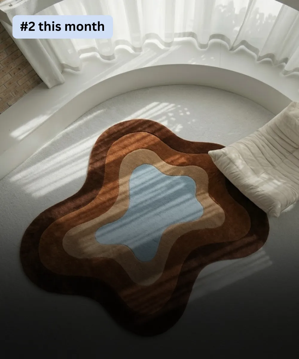 High-angle interior lifestyle shot of a plush irregular rug designed in a fluid, asymmetrical puddle silhouette, styled next to a cream low-profile chair by a bay window with sunbeams highlighting concentric flat 2D color layers of chocolate brown, warm camel, taupe beige, and a sky blue center field.