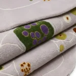 A macro close-up of the folded Liora Moss rug, showcasing the plush, dense pile and the intricate, vibrant green and purple tufted details against a soft grey background.