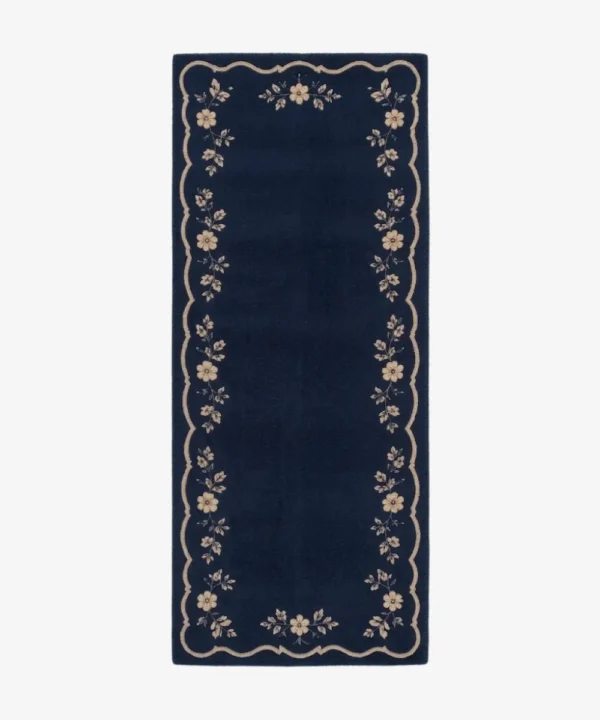 A top-down studio product photograph of a rectangular navy blue runner rug featuring a decorative beige scalloped border and intricate floral vine motifs.