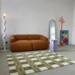 Wide perspective interior lifestyle shot of the Moss Interlace plaid rug styled on a smooth gray concrete floor in a modern room, positioned in front of a plush camel brown sofa and a lavender accent table.