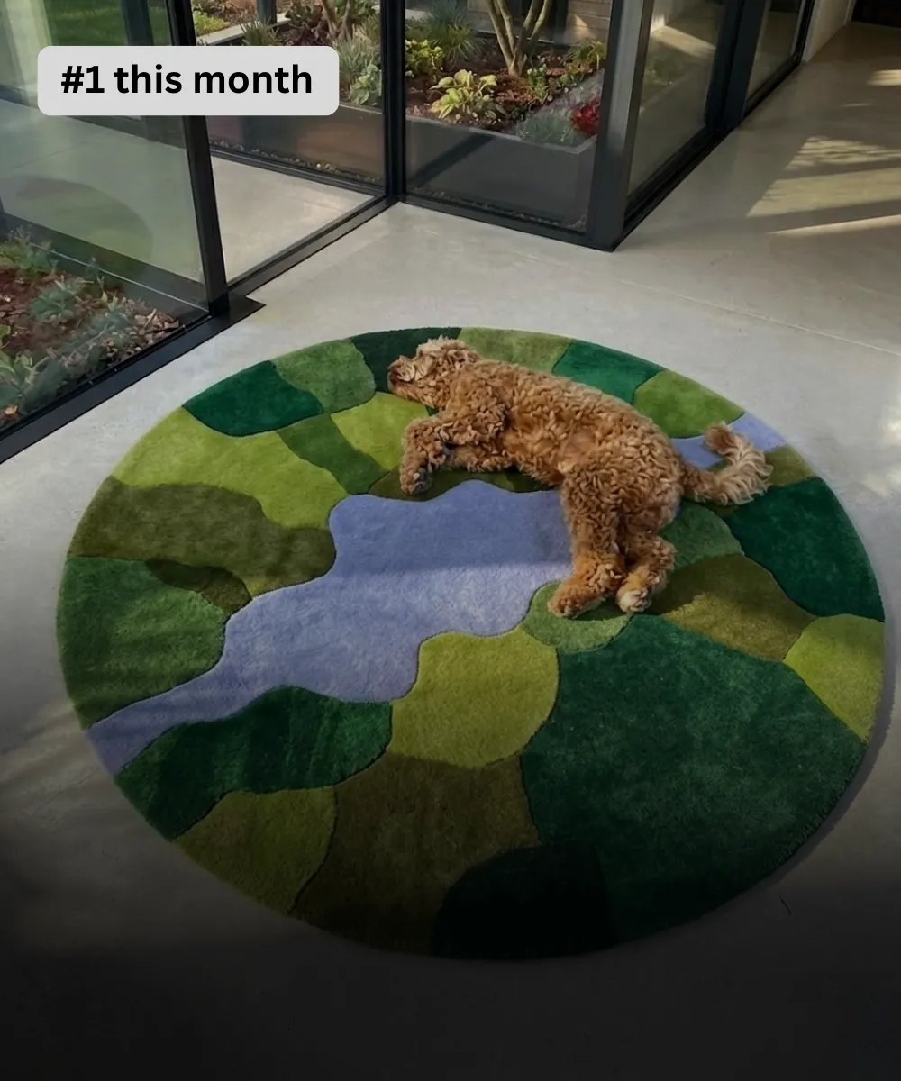 Wide perspective interior lifestyle shot of a round moss rug styled on a concrete floor next to large floor-to-ceiling windows, showcasing an abstract pond design with a lavender blue center water pool surrounded by multi-textured organic shape blocks in forest green, sage, and olive tones while a goldendoodle dog rests on top.
