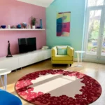 Wide perspective interior lifestyle shot of the circular Rosara red floral rug styled on a light oak hardwood floor in a brightly lit, colorful living room with a pink accent wall and a lime yellow armchair.