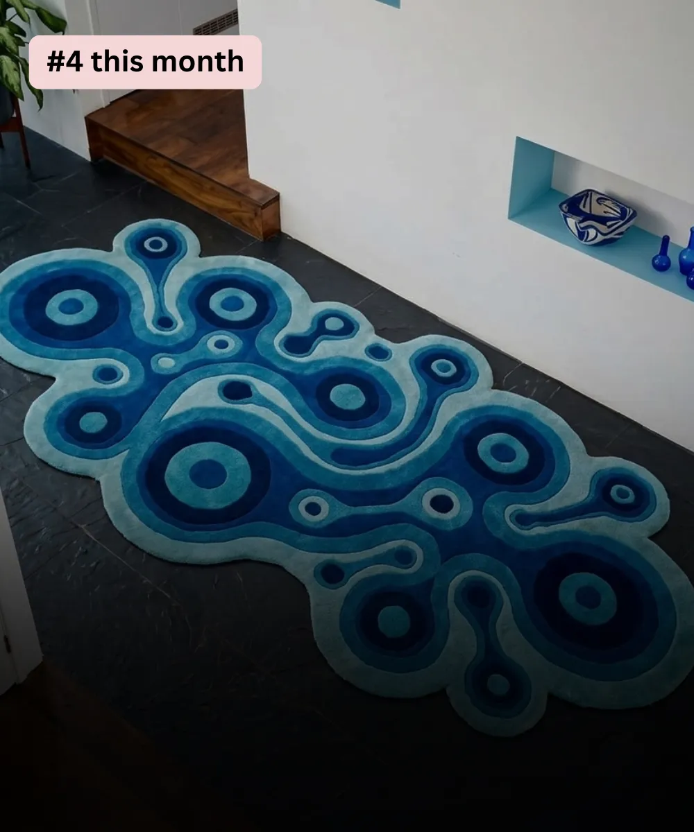 High angle interior lifestyle view of a long runner rug featuring an irregular, wavy silhouette styled on dark slate floor tile, showcasing a flowing psychedelic liquid design composed of concentric target circles and swirling amoeba paths in solid color blocks of navy blue, cyan, and sky blue.