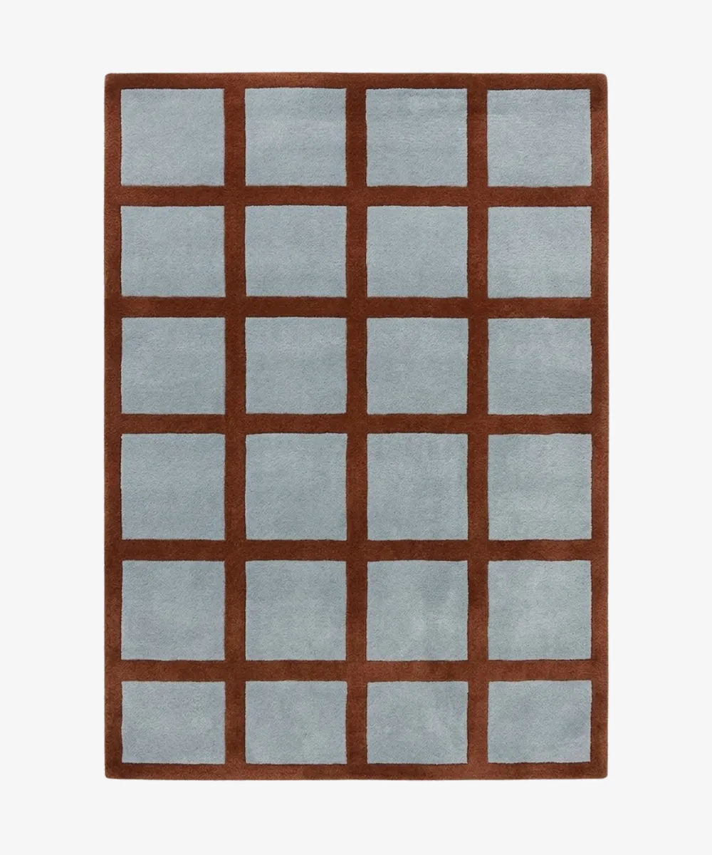 A top-down studio product photograph of the rectangular Russet Ledger area rug, featuring a grid-patterned design with a deep rust-colored border and light blue square inserts.