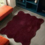 An interior lifestyle shot of a plush deep red scalloped area rug styled on a gray tiled floor in a modern room, positioned near a bold orange bench and blue shelving.