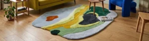 An interior lifestyle photograph of a large, irregularly shaped organic abstract area rug featuring vibrant topographic-style color blocks in green, yellow, blue, and orange on a wooden floor in a modern living room.